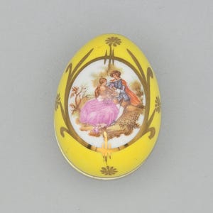 May include: Yellow egg-shaped trinket box with a gold floral design and a central oval scene. The scene depicts a couple in period clothing, with a woman in a pink dress and a man in a blue coat and red cape. The box is likely porcelain.