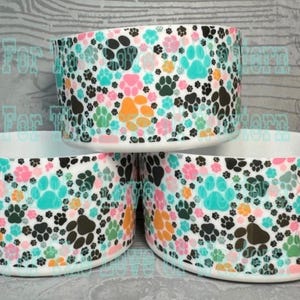 May include: Three white pet bowls with a colorful paw print pattern. The pattern includes black, pink, orange, and teal paw prints on a white background. The bowls are stacked on top of each other.