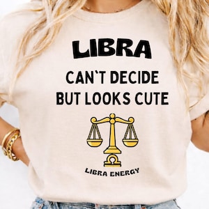 May include: A cream-colored t-shirt with the word "LIBRA" in bold black letters. Below, the text reads "CAN'T DECIDE BUT LOOKS CUTE" with a Libra symbol and "LIBRA ENERGY" in black. The shirt has short sleeves.