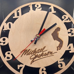 Può includere: Un orologio in legno con numeri neri e sfondo nero. Il quadrante dell'orologio presenta una silhouette di Michael Jackson e la sua firma. Le lancette dell'orologio sono nere e rosse.