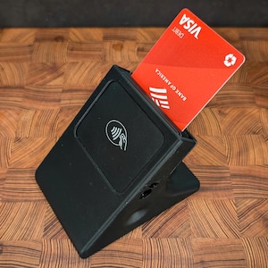 May include: A black plastic card reader with a contactless payment symbol. A red Visa debit card is inserted into the reader. The reader is sitting on a wooden surface.