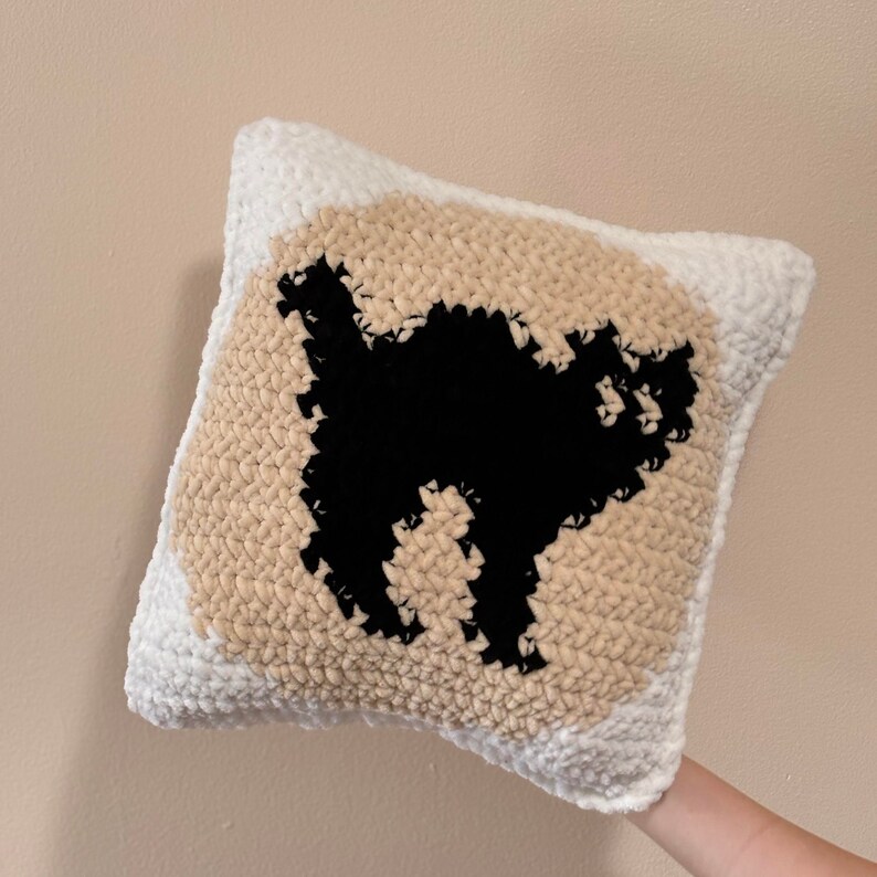 May include: A white and beige throw pillow with a black silhouette of a cat on the front.