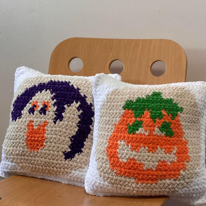 May include: Two white crocheted throw pillows with colorful embroidered designs. One pillow features a purple ghost with orange eyes and the other pillow features an orange pumpkin with a green stem and a jack-o-lantern face.