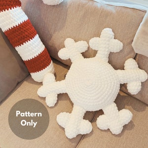May include: White crocheted snowflake pillow with six points. The pillow is on a beige couch with a red and white striped crocheted pillow in the background. The text "Pattern Only" is written in black on a brown circle.