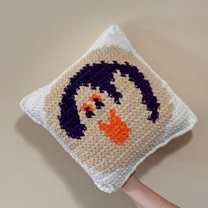 May include: A white crocheted pillow with a beige background and a purple and orange design. The design is a stylized face with a purple mouth and orange eyes.