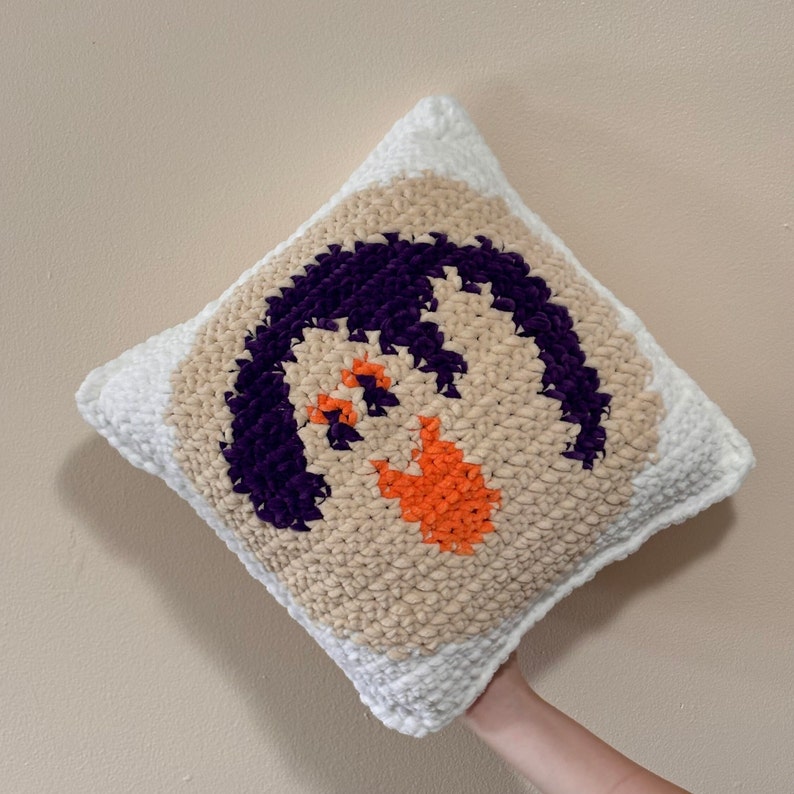 May include: A white crocheted pillow with a beige background and a purple and orange design. The design is a stylized face with a purple mouth and orange eyes.