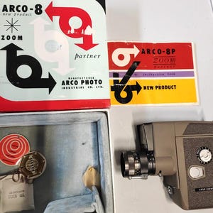 Op de afbeelding: Vintage Arco-8 cameraset met originele verpakking. De set bevat een grijze camera, een instructieboekje en een doos met de tekst "ARCO-8 new product" en "ZOOM". De doos heeft ook de tekst "ARCO PHOTO INDUSTRIAL CO. LTD."