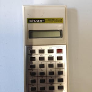 Könnte beinhalten: Ein Vintage Sharp EL-214 Taschenrechner mit cremefarbenem Kunststoffgehäuse. Der Rechner verfügt über einen Bildschirm, eine Tastatur mit Zahlen- und Funktionstasten und das Sharp-Logo. Rechteckige Form.