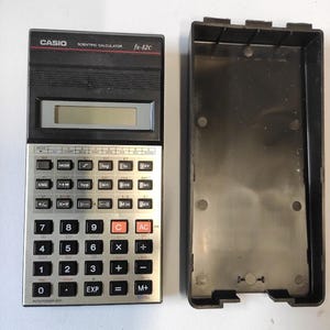 Op de afbeelding: Een zwarte en zilverkleurige Casio fx-82c wetenschappelijke rekenmachine met beschermhoes. De rekenmachine heeft een display, diverse functieknoppen en een numeriek toetsenbord. De hoes is zwart en open, naast de rekenmachine geplaatst.