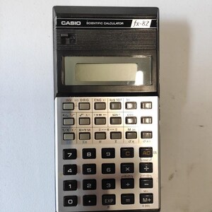 Peut inclure: Calculatrice scientifique Casio fx-82, de couleur noire et argentée. Elle comporte un écran d'affichage et de nombreux boutons pour les fonctions mathématiques. Le nom de la marque et le numéro de modèle sont visibles.