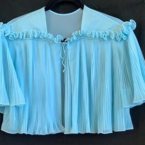 May include: A light blue, pleated, sheer cape with ruffled trim along the neckline and sleeves. The cape has a tie closure at the front and is hanging on a black hanger. The fabric appears lightweight and flowing.