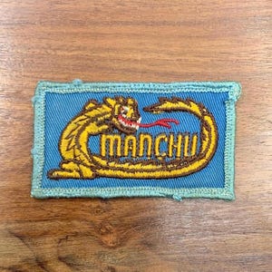May include: A blue embroidered patch with a yellow dragon and the word "MANCHU" in brown lettering.
