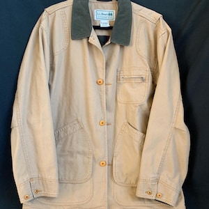 May include: A tan canvas field jacket featuring a dark olive green corduroy collar, a button-front closure with light brown buttons, two large lower patch pockets, and a single zippered chest pocket. The interior label displays the text L.L.Bean.