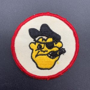 May include: A white embroidered patch with a red border, featuring a cartoon pirate with a black eye patch, black hat, and yellow face holding a cigar.