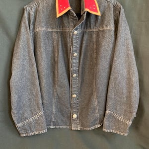 May include: A dark gray denim jacket featuring a vibrant red collar with tan trim and small silver stud accents. The jacket has a button-down front with silver-toned metal buttons, long sleeves with cuffs, and vertical seam detailing on the front.
