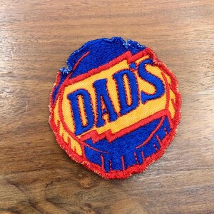 May include: A blue and yellow embroidered patch with the word "DAD'S" in blue lettering.