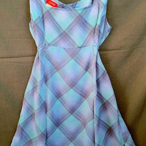 May include: A sleeveless, knee-length dress with a blue and purple plaid pattern. The dress has a square neckline and is hanging on a black hanger. The Esprit brand tag is visible.