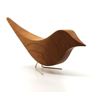 May include: A wooden bird sculpture with a smooth, curved design. The sculpture is made of light brown wood and is supported by two thin, gold-colored metal legs. The bird's body has a curved shape, with a pointed beak and a long, upward-sweeping tail.