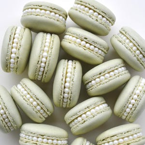 May include: A collection of pale green macarons, each filled with rows of white, pearl-like candies. The macarons are arranged on a white surface, creating a visually appealing display of the sweet treats. The image showcases the delicate texture and color of the desserts.