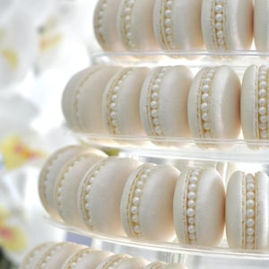 May include: A tiered display of white macarons decorated with pearl-like accents. The macarons are arranged in a pyramid shape on a clear acrylic stand.