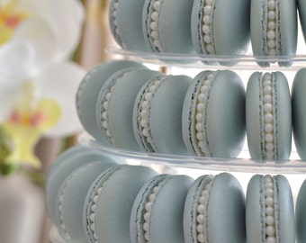 May include: A tiered cake stand with light blue macarons decorated with white pearl-like accents. The macarons are arranged in a circular pattern on each tier.