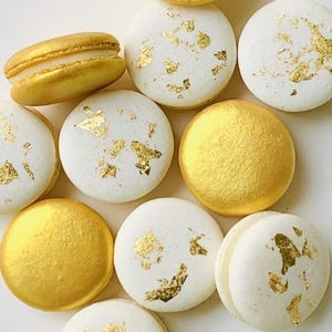 May include: An assortment of macarons, some with a solid gold finish and others with white shells adorned with gold leaf flakes. One macaron is open, revealing a creamy filling. The macarons are arranged on a white surface.