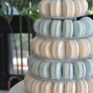 May include: A tower of macarons decorated with pearl-like accents. The macarons are arranged in alternating layers of light blue and ivory. The tower is on a white cake stand with a beaded edge.