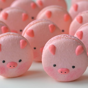 May include: Pink pig-shaped macarons with black eyes and a pink snout. The macarons are arranged in a row on a white surface.