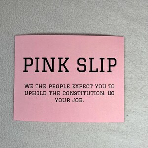 May include: A pink card with the text "PINK SLIP" in large black letters. Below the text is a message that reads "WE THE PEOPLE EXPECT YOU TO UPHOLD THE CONSTITUTION. DO YOUR JOB."