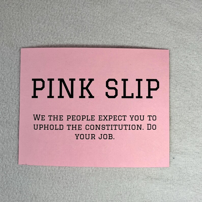 May include: A pink card with the text "PINK SLIP" in large black letters. Below the text is a message that reads "WE THE PEOPLE EXPECT YOU TO UPHOLD THE CONSTITUTION. DO YOUR JOB."