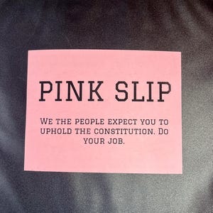 May include: A pink slip of paper with the text "PINK SLIP" in bold black letters. Below the text is a message that reads "WE THE PEOPLE EXPECT YOU TO UPHOLD THE CONSTITUTION. DO YOUR JOB."