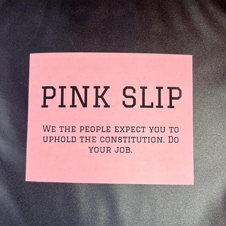 May include: A pink slip of paper with the text "PINK SLIP" in bold black letters. Below the text is a message that reads "WE THE PEOPLE EXPECT YOU TO UPHOLD THE CONSTITUTION. DO YOUR JOB."