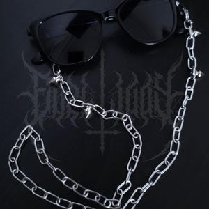 Op de afbeelding: Zwarte zonnebril met een zilveren ketting met conische spikes. De ketting is aan de monturen bevestigd en hangt naar beneden. De zonnebril staat tegen een donkere achtergrond met een gotisch design.