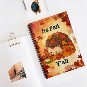 Può includere: Un taccuino a spirale con un riccio dei cartoni animati circondato da foglie autunnali e un fungo. La copertina riporta la scritta "Its Fall Y'ALL" in caratteri neri su uno sfondo dai toni arancioni caldi con foglie d'acero sparse.