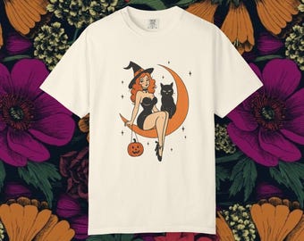 May include: Cream-colored t-shirt featuring a retro illustration of a witch with red hair, a black cat, and a jack-o'-lantern. The witch is seated on an orange crescent moon. The design has a vintage Halloween theme.