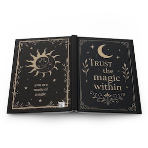May include: An open hardcover book with a black cover and gold-colored illustrations. The left page features a sun and moon design with the text "you are made of magic". The right page displays a crescent moon and the text "Trust the magic within".