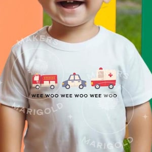 Può includere: Una maglietta bianca per bambini con un camion dei pompieri rosso, un'auto della polizia blu e un'ambulanza bianca con una croce rossa. Il testo "WEE WOO WEE WOO WEE WOO" è stampato sotto i veicoli.