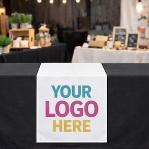 May include: A black table is covered with a white table runner. The runner features the text "YOUR LOGO HERE" in teal, pink, and yellow. The text "Option 2: 24" x 72"" is at the top of the image. The background is blurred.