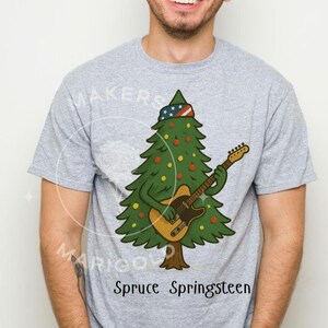 May include: Light gray t-shirt featuring a cartoon Christmas tree playing a guitar. The tree wears a hat with a stars and stripes design and is decorated with ornaments. The text "Spruce Springsteen" is printed below the graphic.