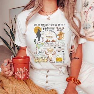 May include: White t-shirt with a country music themed design. The design includes a cowboy riding a horse, a guitar, a margarita glass, a skull, a beer bottle, and the text "What Makes You Country", "Play It Again", "Country Girl", "Sunrise Sunburn Sunset", "Rollecoaster", "Do I", "Drink A Beer", "I Don't Want This Night To End", "Kick The Dust Up", and "Drunk On You".
