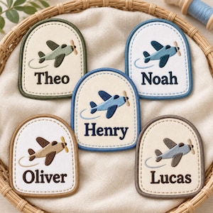 May include: Five arched fabric patches with embroidered airplane motifs and names. The patches feature the names Theo, Noah, Henry, Oliver, and Lucas in dark thread, each accompanied by a small airplane graphic with a propeller and a flight path line.