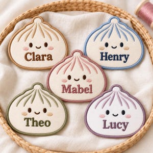 May include: Five embroidered patches shaped like smiling dumplings, each featuring a unique colored border and text. The names Clara, Henry, Mabel, Theo, and Lucy are stitched in matching thread colors on cream-colored fabric with pink blush details.