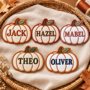 May include: Five embroidered pumpkin-shaped patches with cream-colored fabric and brown stems with green leaves. Each patch features a name stitched in the center: Jack in orange, Hazel in brown, Mabel in pink, Theo in green, and Oliver in blue.