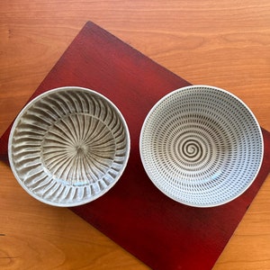 May include: Two ceramic bowls with unique designs. One bowl features a radial pattern in shades of brown, while the other has a spiral pattern in black and white. The bowls are placed on a red surface, with a wooden background.
