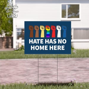 May include: A dark blue yard sign with the text "HATE HAS NO HOME HERE" in white. Above the text are illustrations of fists in various colors, including brown, yellow, red, and rainbow. The sign is displayed outdoors.