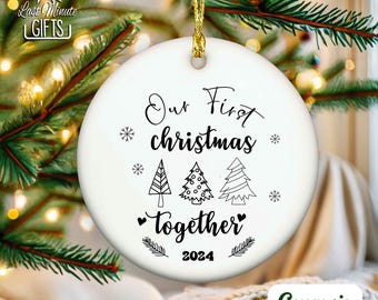 May include: A round white ceramic ornament featuring black text that reads Our First Christmas together 2024. The design includes three stylized line-art Christmas trees, small snowflakes, and two hearts, suspended by a gold string.