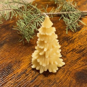 Puede incluir: Una vela en forma de árbol de color amarillo claro con una superficie texturizada. La vela está hecha para parecerse a un árbol de Navidad y se coloca sobre una superficie de madera. Una ramita de agujas de pino verdes está en el fondo.
