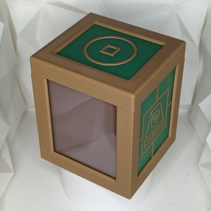 May include: A brown wooden box with a green top. The top has a gold circle with a square inside. The box has a clear window on the front.