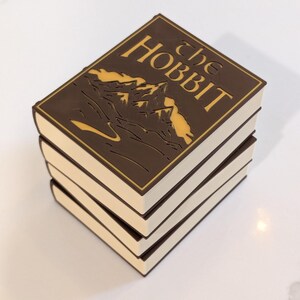 May include: A stack of three rectangular objects resembling books with brown covers and white edges. The top cover features a gold-colored mountain graphic and the text "the HOBBIT" in a stylized font, enclosed within a thin gold rectangular border.