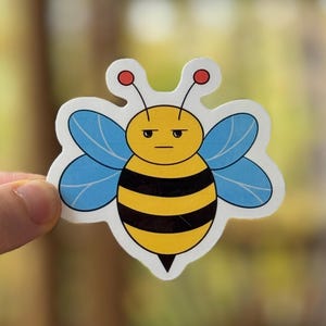 May include: A die-cut sticker featuring a cartoon bee with a grumpy expression. The bee has a yellow body with black stripes, blue wings, and red antennae. The sticker is held by a hand against a blurred background.